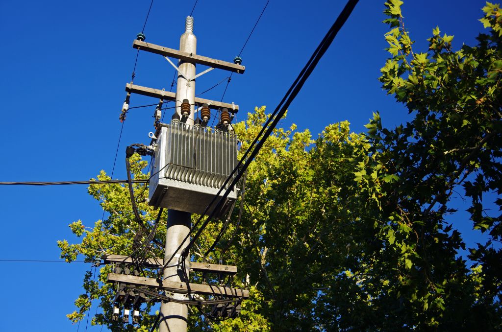 Power transformer on a concrete electricity pole against tree canopies and a clear blue sky