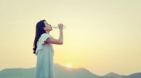 Side View Of Young Woman Drinking Water While Standing Against Sky During Sunset