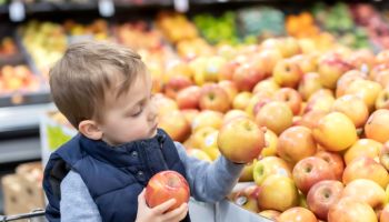 Three years old little child sitting in a shopping cart buying apples at a supermarket aisle