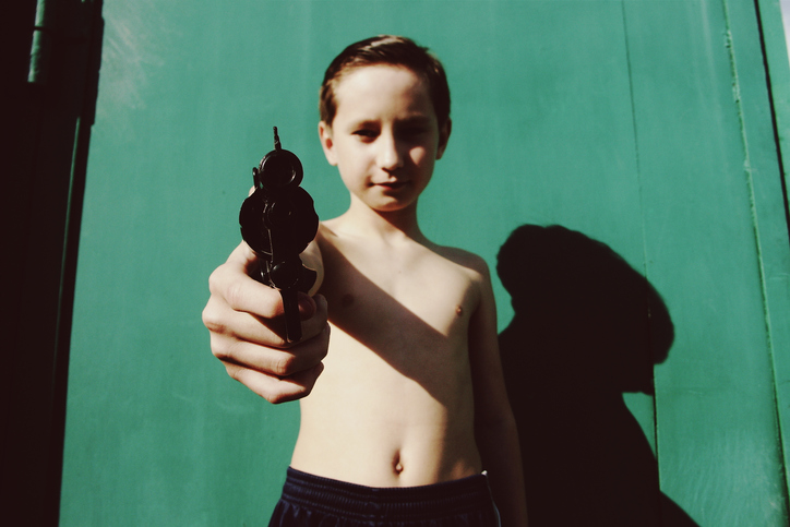 Portrait Of Shirtless Boy Holding Toy Gun Against Green Wall During Sunny Day