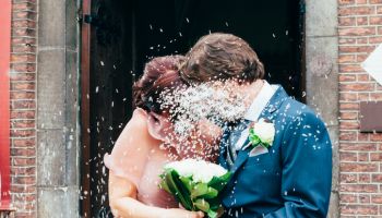 Petals Falling On Groom And Bride With Bouquet Standing Outside Building