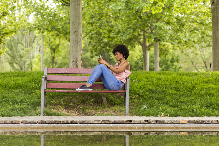 Smiling Woman Using Mobile Phone While Sitting On Wooden Bench At Park
