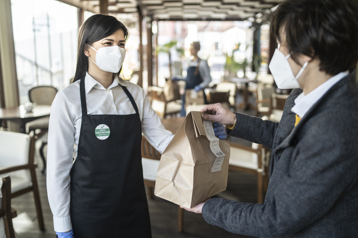 Restaurant employee wearing a sticker showing that she has been vaccinated against COVID-19, giving a take away food to a customer.