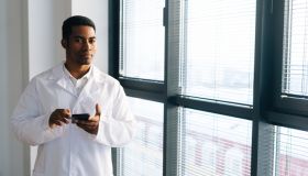 Serious black male doctor wearing white uniform coat using typing on smartphone standing by window at hospital office.