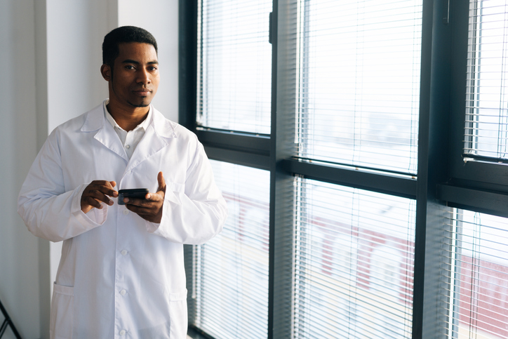 Serious black male doctor wearing white uniform coat using typing on smartphone standing by window at hospital office.