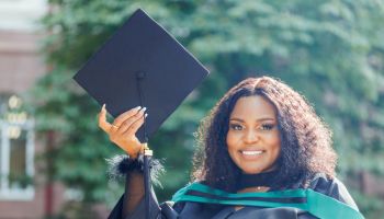 Happy black female graduate with natural curly black hair in graduation gown holding mortarboard and smiling