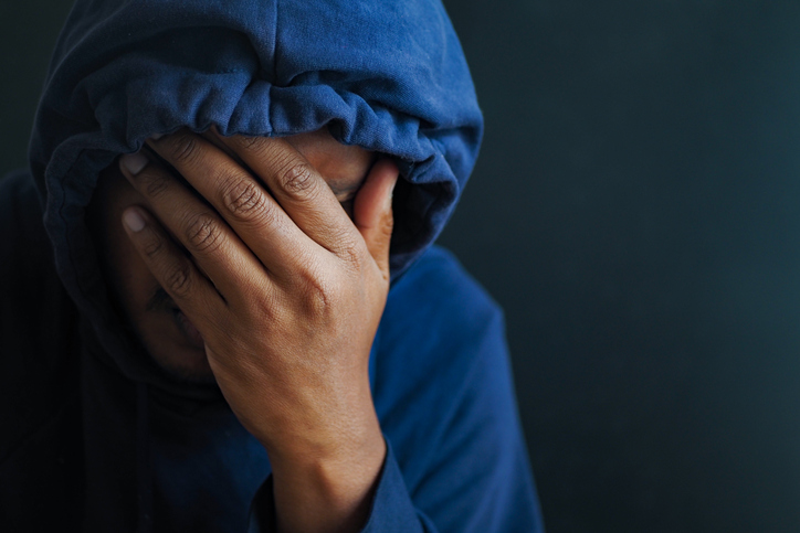 sad man in hood cover face with hands isolated in black.
