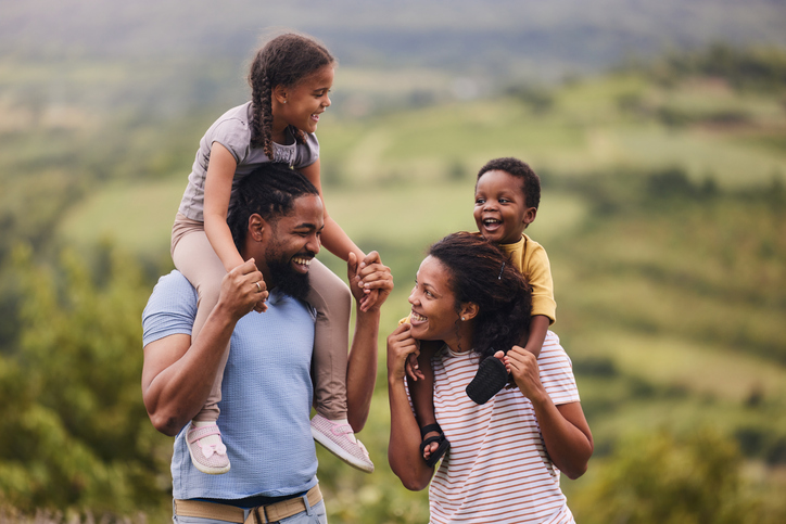 Happy black parents carrying their small kids on shoulders in nature.