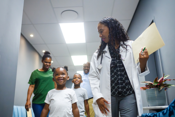 Female doctor walking with brave young cute girl in hospital corridor to consultation with family following
