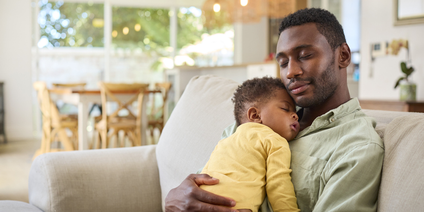 Family Shot With Father Cuddling Baby Daughter Sleeping On Sofa At Home Together